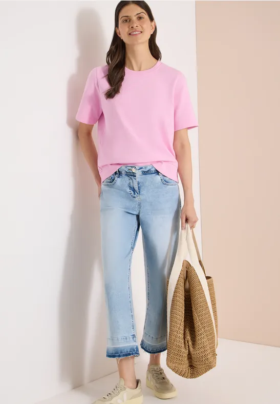 Eine Person trägt ein rosafarbenes T-Shirt, hellblaue Jeans und hält eine geflochtene Tasche, dazu Sneaker.