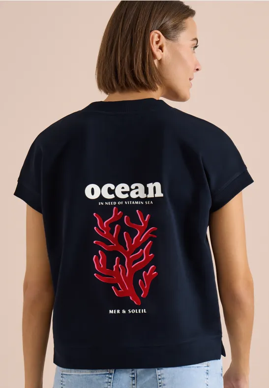 Frau trägt dunkelblaues T-Shirt mit weißem 'ocean'-Schriftzug und rotem Korallenprint auf dem Rücken.