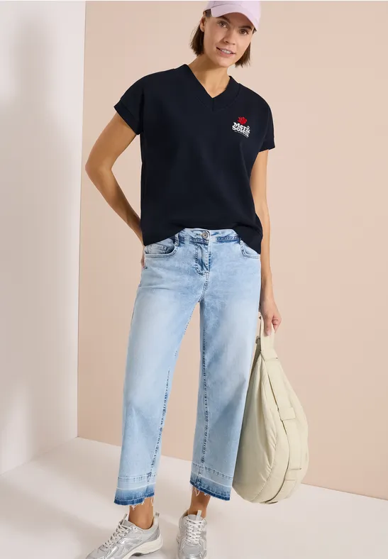 Frau in schwarzem T-Shirt, hellblauen Jeans und Shopper-Tasche vor hellem Hintergrund.