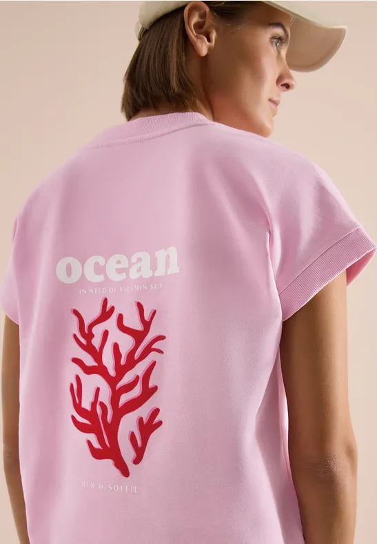 Eine Person trägt ein rosa T-Shirt mit dem Schriftzug „ocean“ und einer roten Koralle auf dem Rücken.