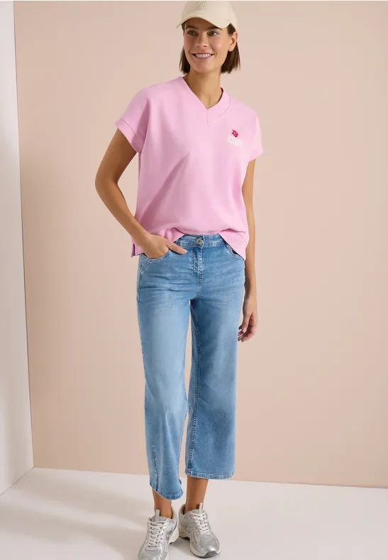 Frau trägt pinkes T-Shirt, hellblaue, weite Cropped-Jeans und weiße Sneaker vor beigem Hintergrund.