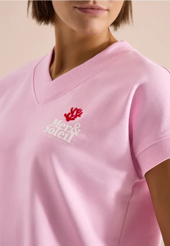 Frau trägt rosa V-Neck-Shirt mit rotem Korallensymbol und weißem Schriftzug.
