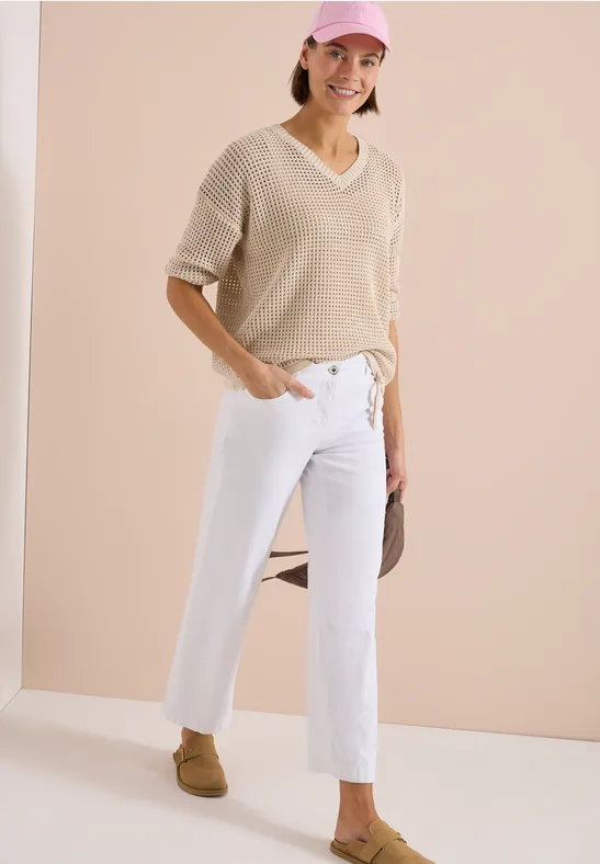 Een persoon draagt een beige V-hals trui, witte jeans en bruine loafers.