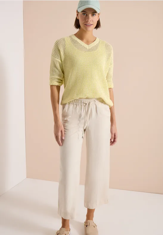 Vrouw met gele trui en lichte, losse broek voor een rustige achtergrond.