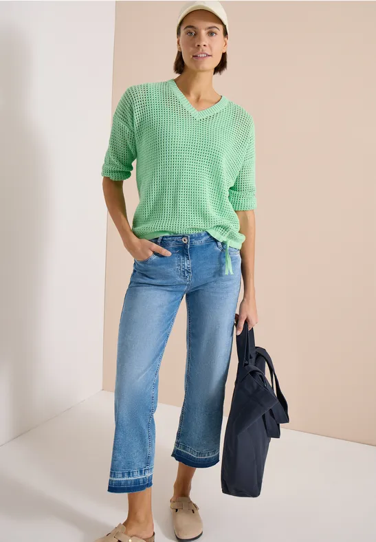 Vrouw in groene trui, cropped wijdere jeans en schoenen, houdt donker colbert vast tegen een neutrale achtergrond.