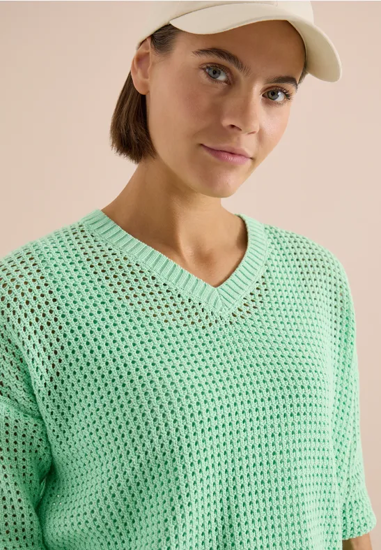 Een persoon met een lichtgroene, losgehaakte V-hals trui en een pet voor een beige achtergrond.