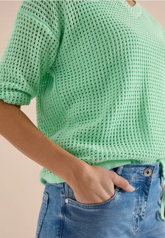 Persoon draagt een mintgroene, losgebreide V-hals trui, hand in de zak van een blauwe spijkerbroek.