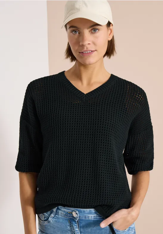 Persoon met zwarte, losse gebreide trui met V-hals en driekwartmouwen, hand in spijkerbroekzak.