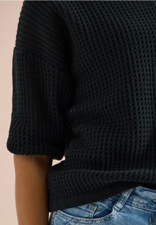 Bovenlichaam gekleed in een zwart grofgebreide trui met driekwartmouwen en een blauwe spijkerbroek.