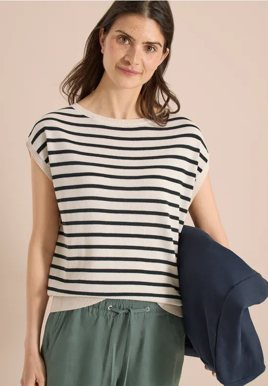 Frau trägt gestreiftes Top, grüne Hose und hält eine dunkelblaue Jacke.