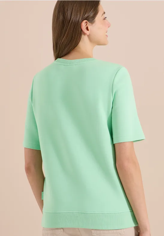 Rückansicht einer Person mit mintgrünem T-Shirt und halblangem Haar vor hellem Hintergrund.