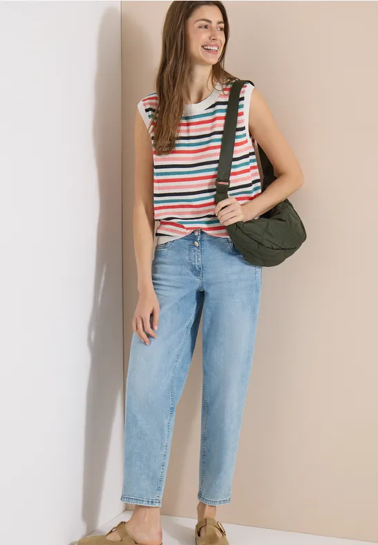 Frau in gestreiftem ärmellosem Top, hellblauen Jeans und grüner Umhängetasche vor beiger Wand.