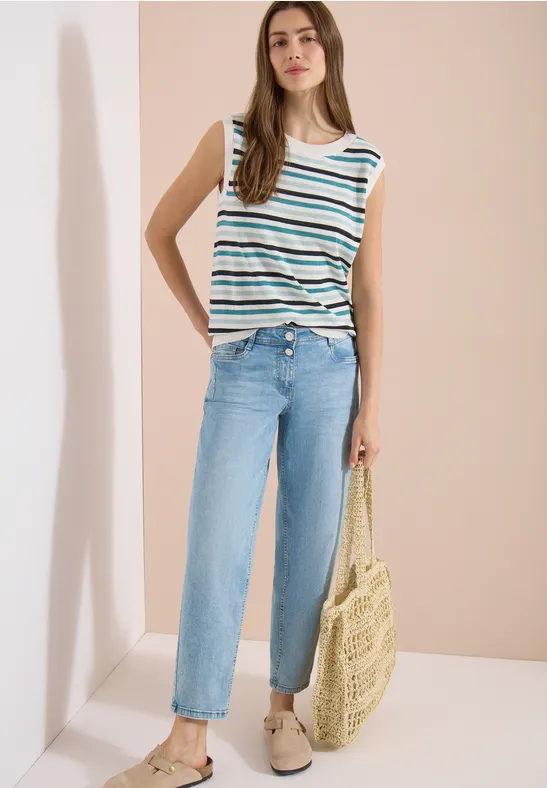 Frau in gestreiftem Top, hellblauen Jeans und Basttasche vor neutralem Hintergrund.