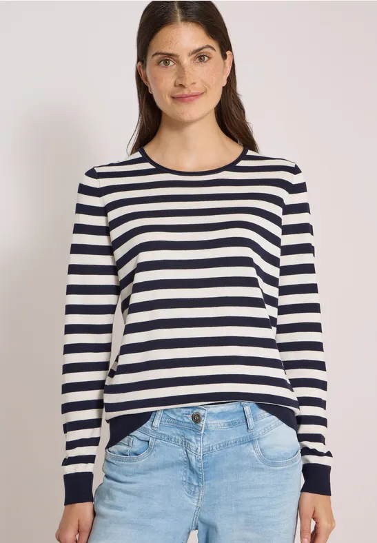 Junge Frau in gestreiftem Langarmshirt und Jeans, lächelt vor weißem Hintergrund.