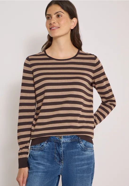 Frau mit braun-schwarz gestreiftem Pullover und blauen Jeans, halb lächelnd vor hellem Hintergrund.