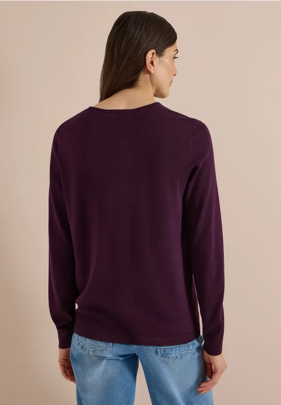Frau von hinten in lila Pullover und Jeans vor neutralem Hintergrund.