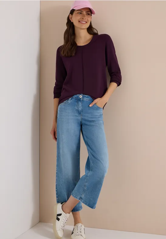 Lachende Frau in lila Top, pinker Cap, weiten hellblauen Jeans und weißen Sneakern lehnt an Wand.