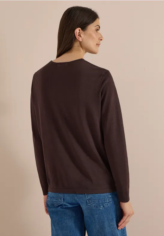 Une femme de profil porte un pull brun foncé simple et un jean bleu devant un fond clair.