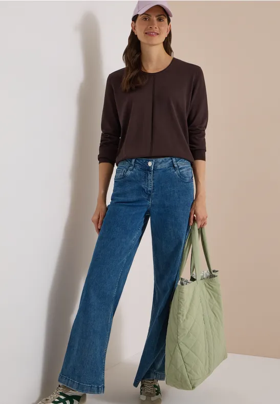 Femme en haut noir à manches longues, jean bleu évasé fendu devant et sac matelassé vert clair.