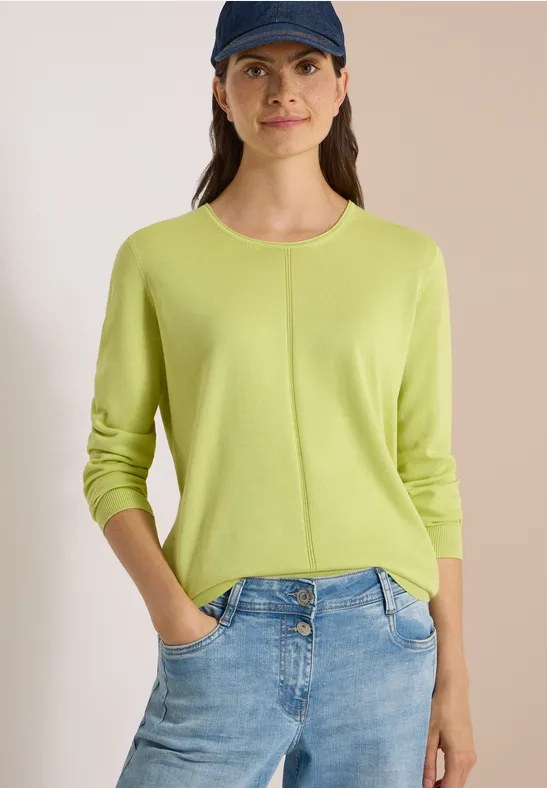 Femme en pull vert citron et jean clair, mains dans les poches, coiffée d'une casquette sombre.
