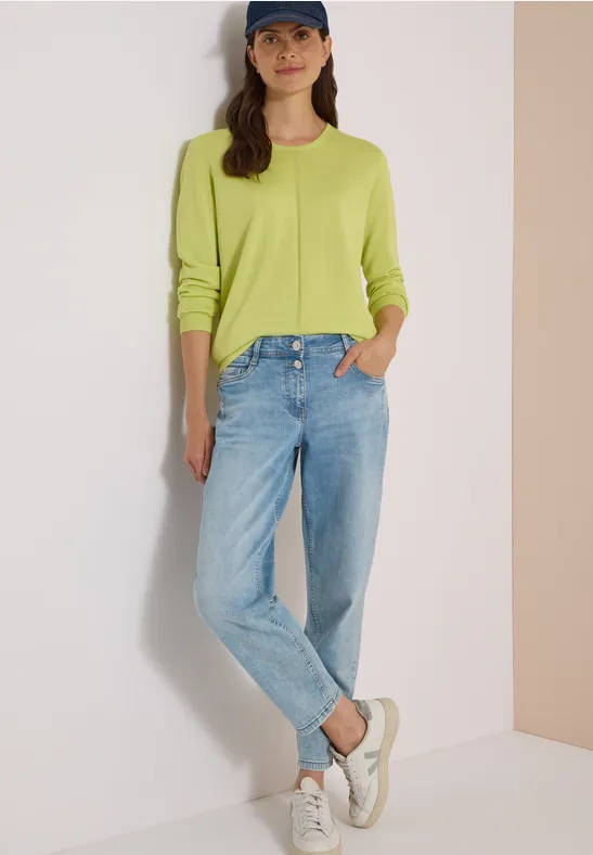 Une femme porte un pull vert vif, un jean bleu, des baskets blanches et une casquette sur fond clair.
