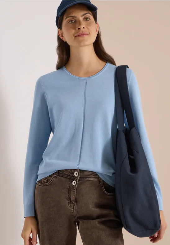Femme en pull bleu clair, pantalon marron, grand sac bleu et casquette.