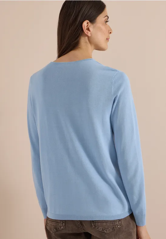 Femme de dos portant un pull bleu clair et un pantalon foncé sur fond neutre.