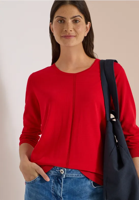 Femme souriante en pull rouge et jean, main dans la poche, avec un sac à bandoulière.