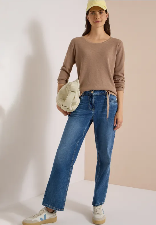Frau trägt Beige-Pullover, blaue Jeans, weiße Sneakers, Cap und Tasche, steht vor neutralem Hintergrund.