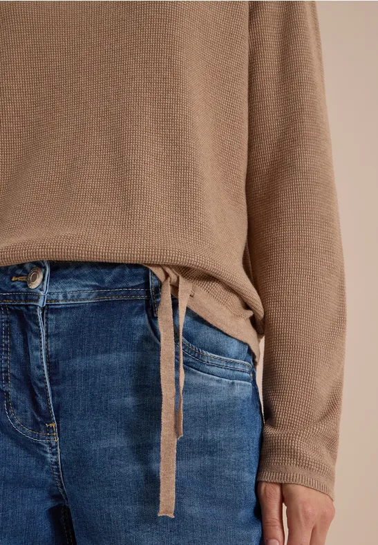 Eine Person trägt einen hellbraunen Pullover mit seitlicher Bindeschleife und blaue Jeans.