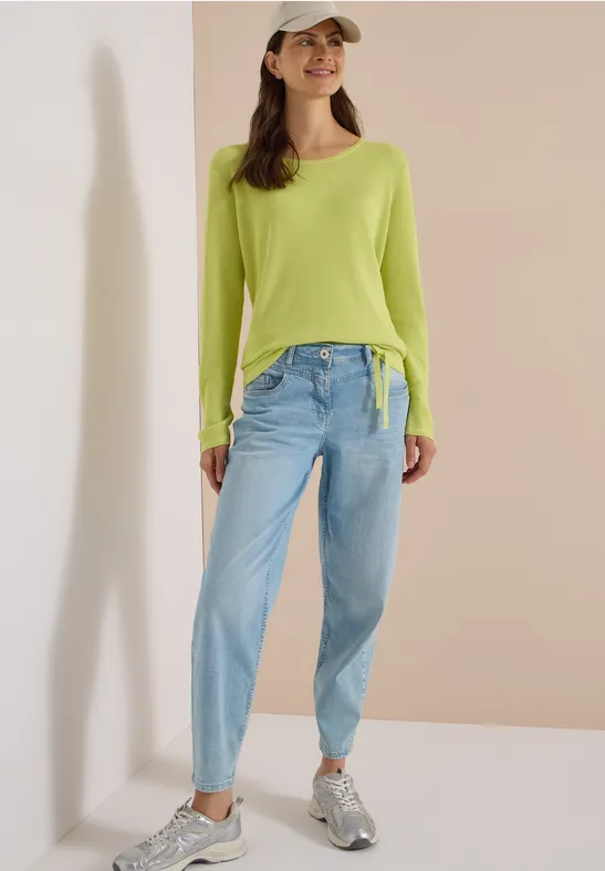 Frau trägt hellgrünes langärmeliges Top, hellblaue Jeans, graue Sneaker und Cap vor neutraler Wand.