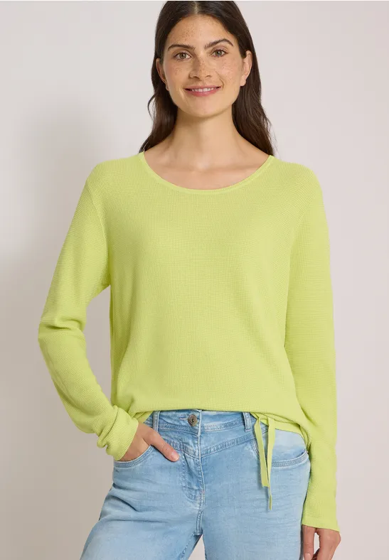 Frau trägt hellgrünen Pullover mit Tunnelzug am Bund und blaue Jeans, lächelt vor neutralem Hintergrund.