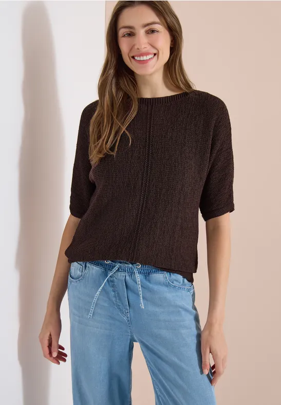 Eine Person trägt einen braunen Strickpullover mit halblangen Ärmeln und hellblaue Jeans.