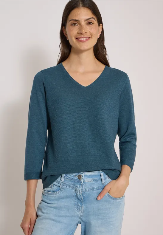 Frau mit grünem V-Ausschnittpullover (3/4-Ärmel) und Jeans, lächelnd, eine Hand in der Hosentasche.