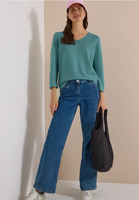 Frau in weiten blauen Jeans, grünem Pullover und rosa Cap, hält eine schwarze Tasche.