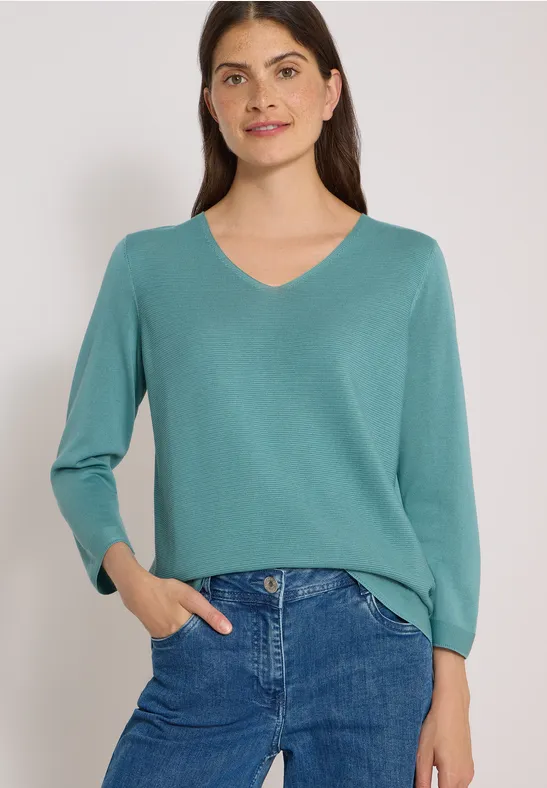 Frau in türkisfarbener V-Kragen-Pullover mit blauer Jeans, lächelnd, eine Hand in der Tasche.
