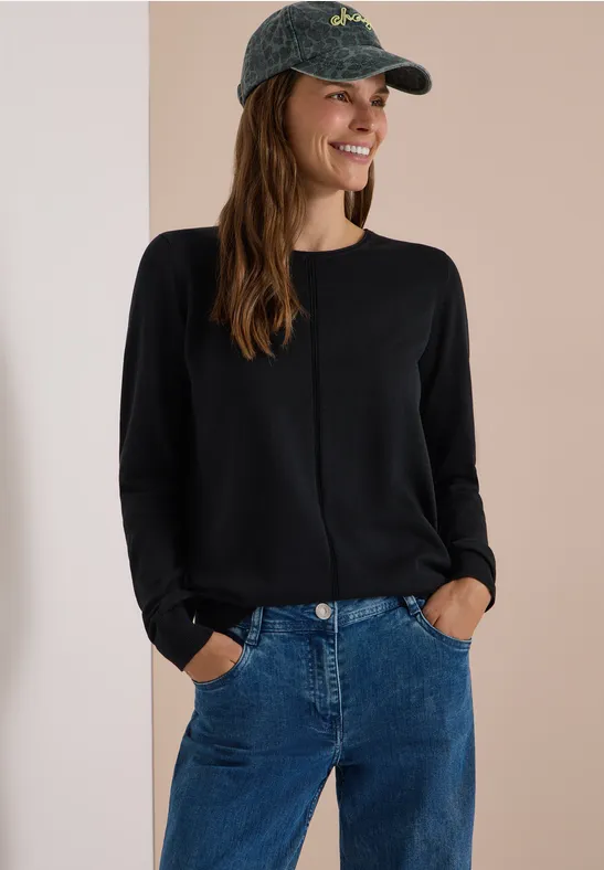 Frau im schwarzen Langarmshirt, blauen Jeans und grüner Baseballkappe, die lächelnd in die Kamera schaut.