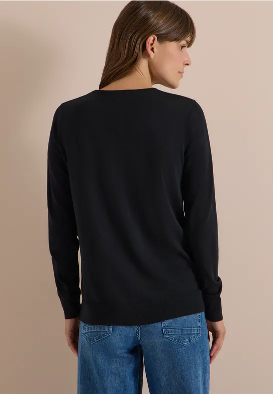 Eine Frau steht mit dem Rücken zur Kamera, trägt schwarzen Pullover und blaue Jeans vor hellbeigem Hintergrund.