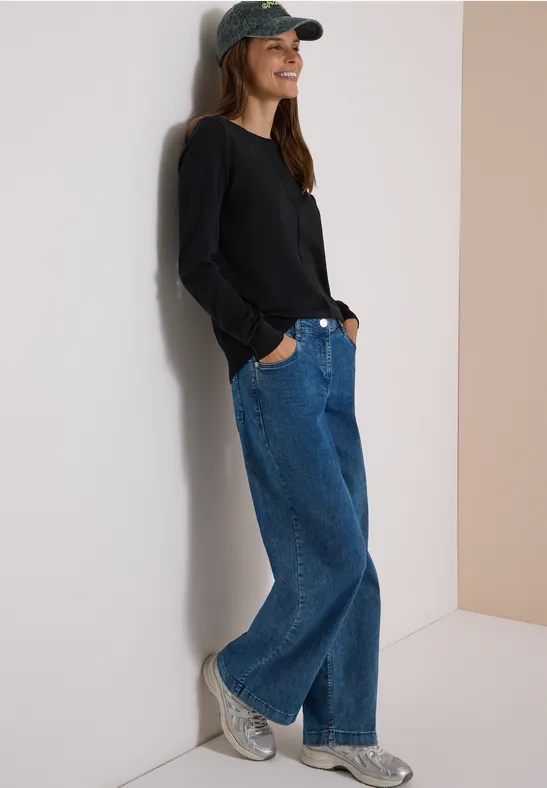Frau mit Baseballcap, schwarzem Pullover und weiten Jeans lehnt lässig an der Wand und lächelt.