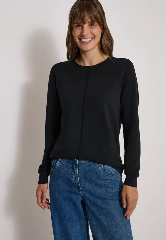Frau mit schwarzem Langarmshirt und blauen Jeans, die lächelnd in die Kamera schaut.