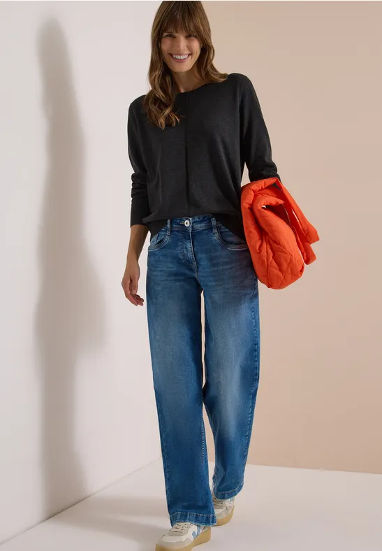 Femme souriante en tee-shirt noir à manches longues et jean bleu tenant une doudoune orange sous le bras.