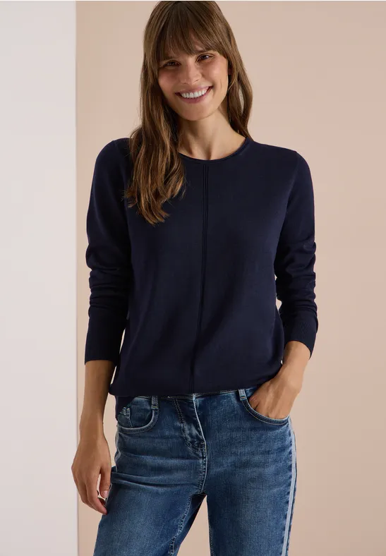 Eine lächelnde Frau trägt einen dunkelblauen Pullover und Jeans.