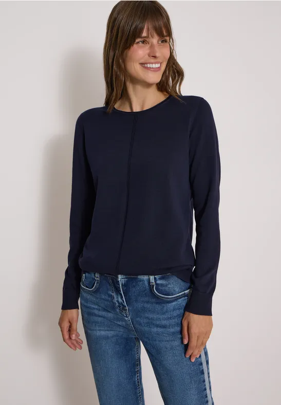 Frau in dunkelblauem Pullover und Jeans steht lächelnd vor hellem Hintergrund.