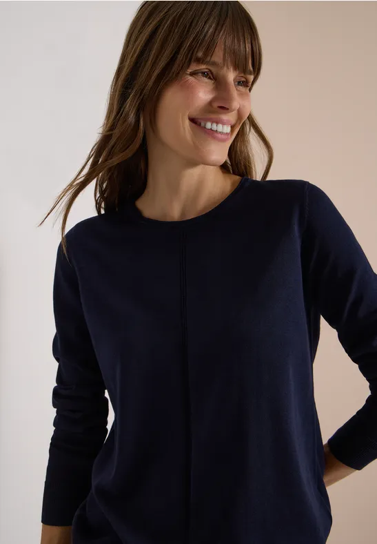 Eine lächelnde Frau trägt einen dunkelblauen Feinstrickpullover, steht entspannt mit Händen an der Hüfte.