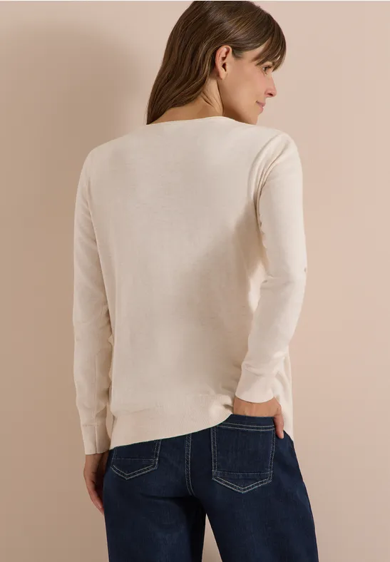 Eine Frau von hinten in einem hellbeigen Langarmpullover und blauer Jeans vor hellem Hintergrund.