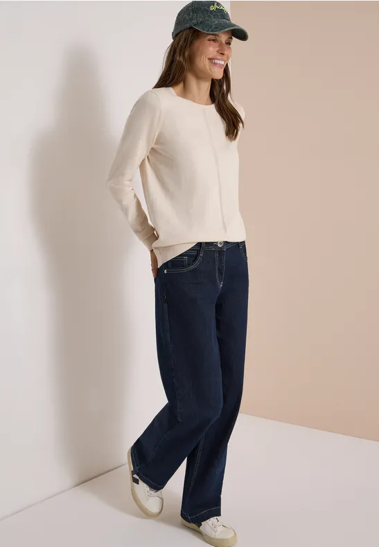 Frau in beigem Pullover, breiten Jeans, weißen Sneakern und grüner Baseballkappe lehnt lächelnd in der Ecke.