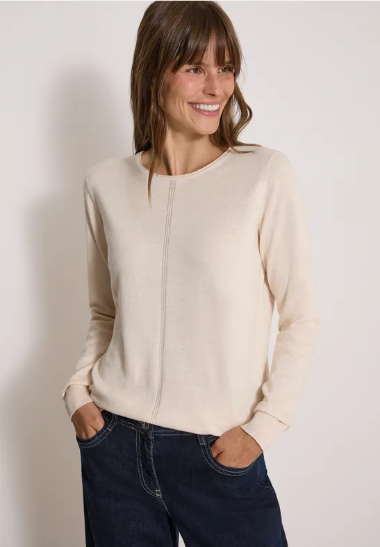 Eine lächelnde Frau trägt einen hellbeigen Pullover und blaue Jeans vor hellem Hintergrund.