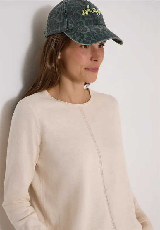 Eine junge Frau trägt einen hellbeigen Pullover mit mittiger Naht und eine dunkelgrüne Cap mit Muster.
