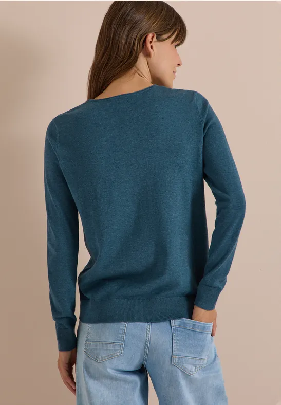 Frau von hinten in petrolfarbenem Pullover und hellblauen Jeans vor neutralem Hintergrund.
