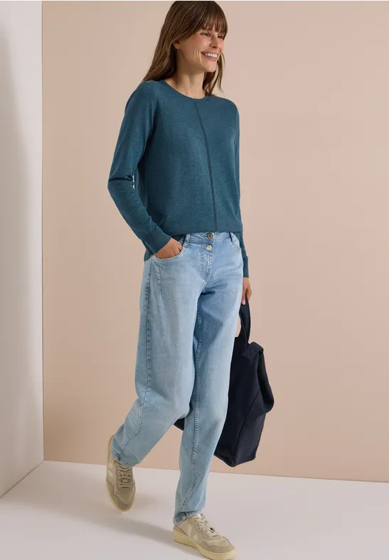 Eine Frau in locker sitzender Jeans, tealfarbenem Pullover und hellen Sneakern, die lächelnd eine Jacke hält.